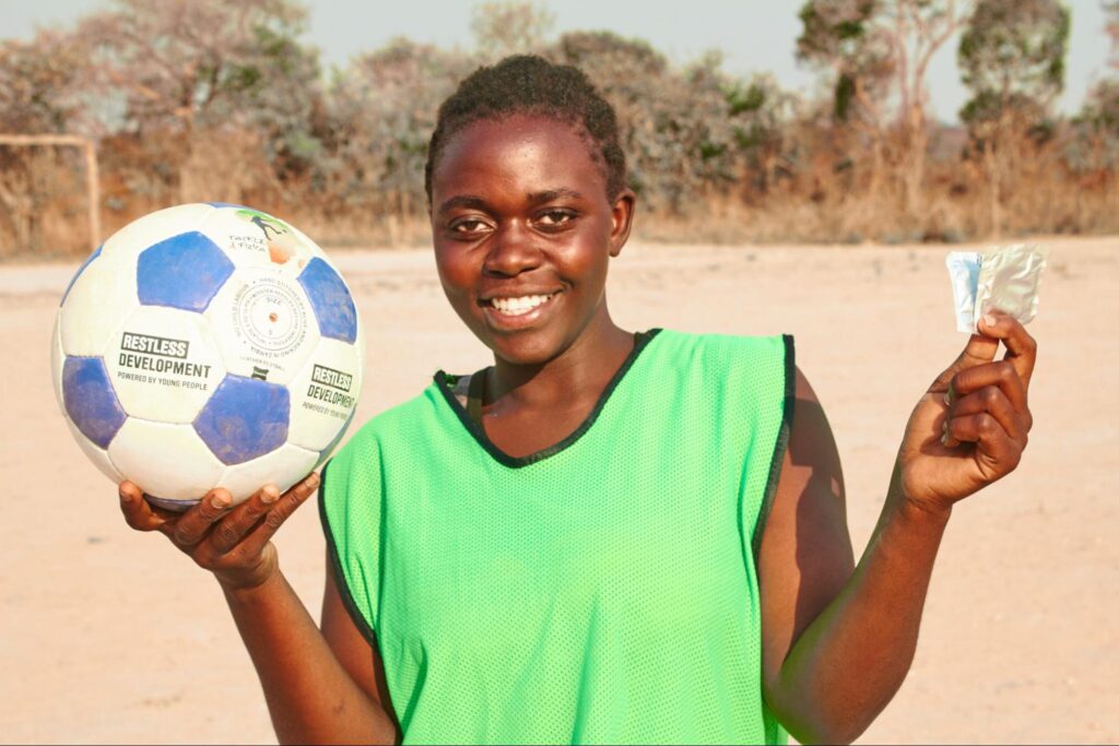 Agness poses for the camera with a soccer ball on her right hand and two packets on condom on the other. 